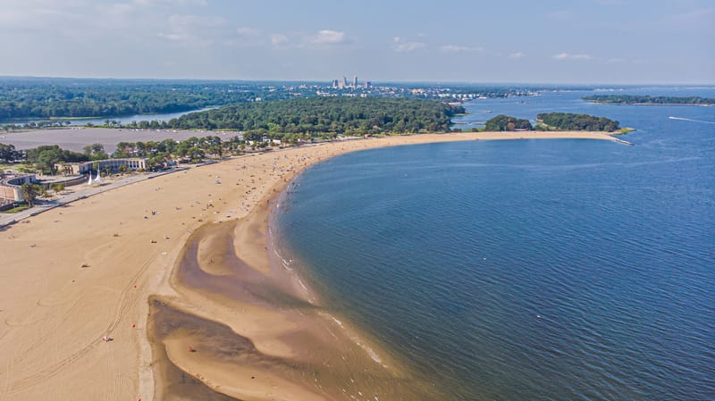 Aerial view of the curved sand of Orchard Beach beside the blue water of Pelham Bay